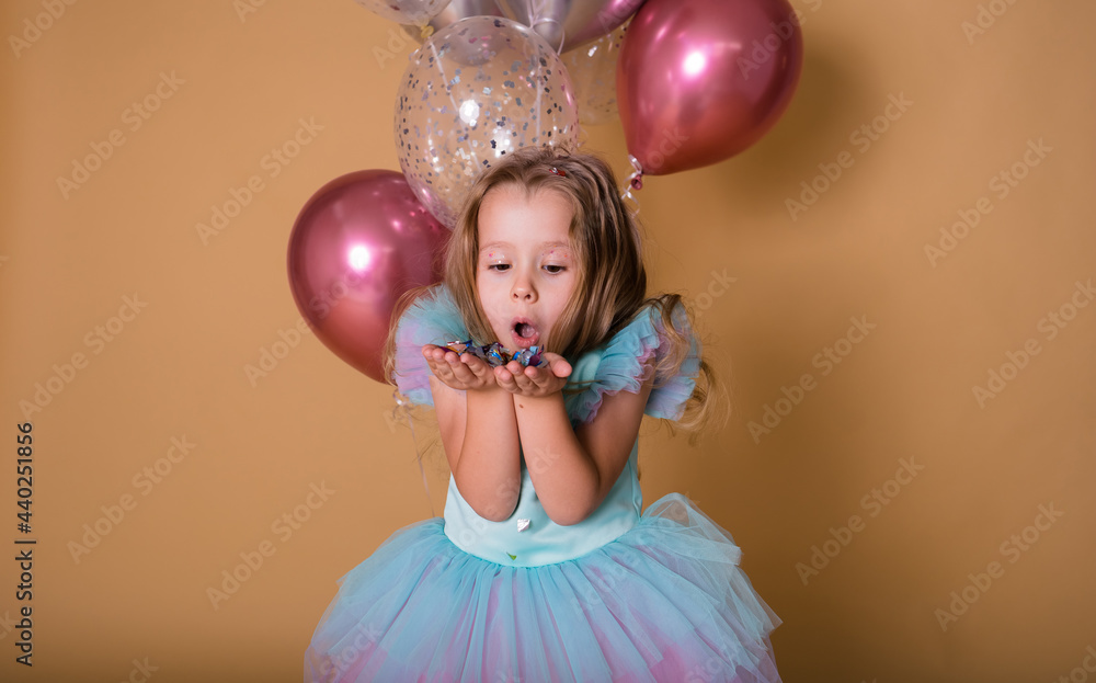 ittle girl in a festive dress plays with colorful confetti on a beige background with balloons. Festive background