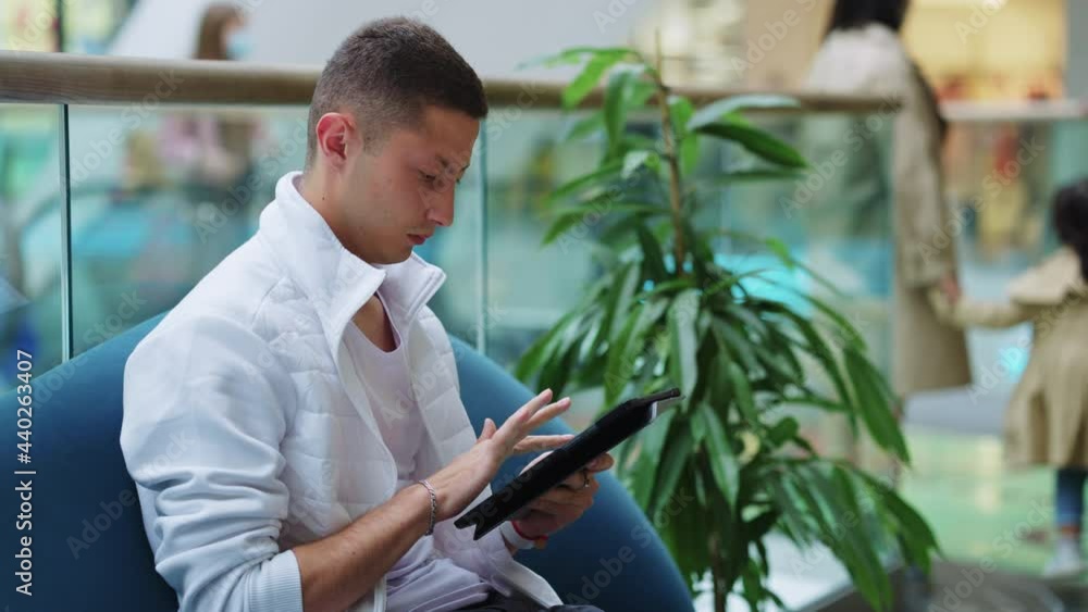 Young man wearing stylish outfit sitting on sofa in mall and using tablet, blurred woman holding child by hand on blurred background, people wearing masks. Concept of gadget Young man wearing stylish outfit sitting on sofa in mall and using tablet, blurred woman holding child by hand on blurred background, people wearing masks. Concept of gadget