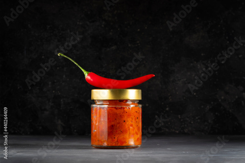 Spicy homemade harissa or adjika sauce of sweet peppers, cumin, coriander and chilli peppers in a glass jar on a dark background photo