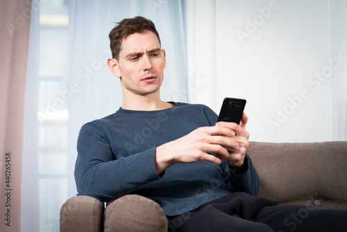 Portrait of sad unpleasant unhappy guy, young frowning man looking at screen of his cell mobile phone, using smartphone with negative face, sitting at home in living room on sofa or couch. Bad news photo