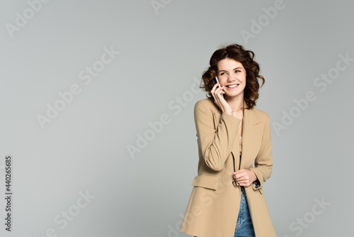 happy woman in beige blazer talking on smartphone isolated on grey
