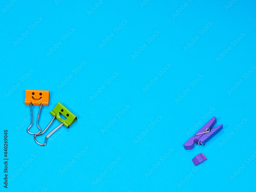 Orange and green metal binder clip or paperclip on blue background with copyspace for text. Different, stand out of the crowd concept, deride. Spring-type wooden violet clothespin with fracture leg Orange and green metal binder clip or paperclip on blue background with copyspace for text. Different, stand out of the crowd concept, deride. Spring-type wooden violet clothespin with fracture leg
