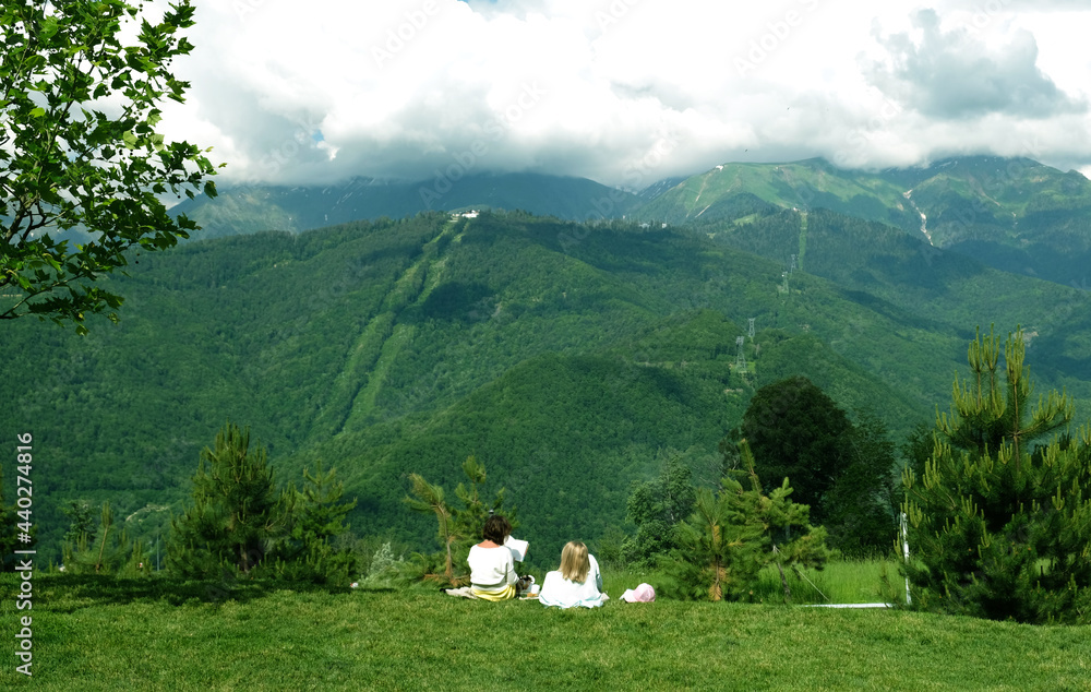Two young women in white sit and lie on the green grass and read books against the backdrop of mountains. Relaxing moment. Beautiful mountain landscape, mountain peak in the clouds.