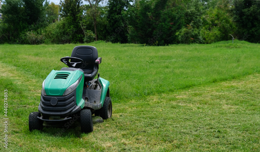 Green grass trimming with lawn mower. Tractor mowing grass in a public park. A dirty lawn tractor after mowing the lawn stands on the meadow. Professional lawn mower cuts the grass. Green grass trimming with lawn mower. Tractor mowing grass in a public park. A dirty lawn tractor after mowing the lawn stands on the meadow. Professional lawn mower cuts the grass.