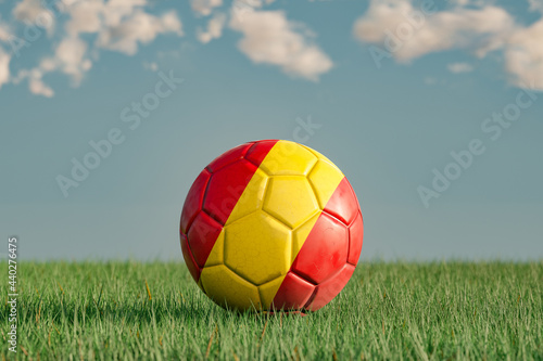 Spanish football with the national colors of Spain on a green meadow. Leather in slightly used look. Background blue with clouds. 3D illustration.
