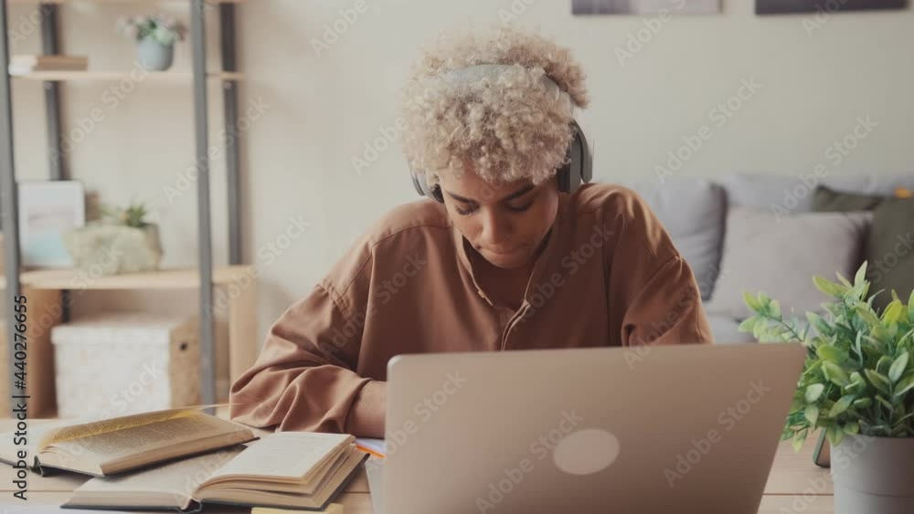 African American female student wear wireless headphone study online with skype teacher learn language listen lecture watch webinar write notes look at laptop sit in home office distance education African American female student wear wireless headphone study online with skype teacher learn language listen lecture watch webinar write notes look at laptop sit in home office distance education