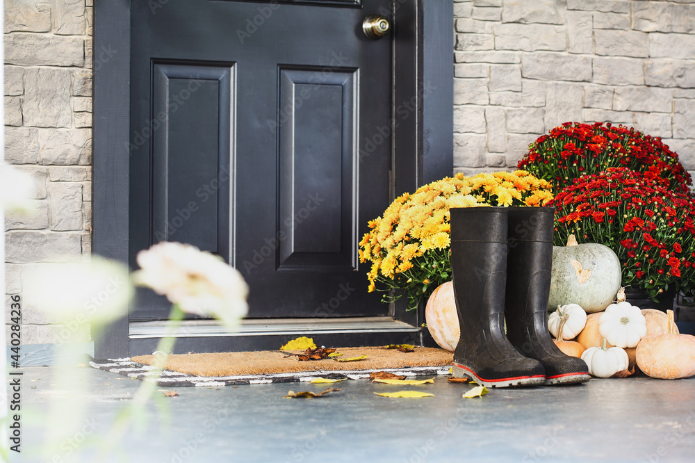 Rain boots sitting on door mat of front porch that has been decorated for  autumn with heirloom white, orange and grey pumpkins and mums. Selective  focus with blurred foreground and background. Stock