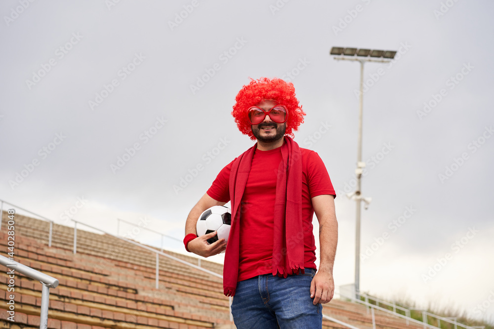 Soccer fan with wig and clown glasses in the stadium looking at the camera and holding a ball.