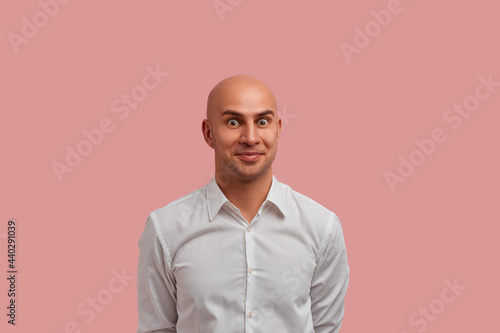 And what next. Pleasantly shocked bald guy in white shirt holds hands behind back. Keeps eyes widely opened, looks what happens next. Isolated over pink background.