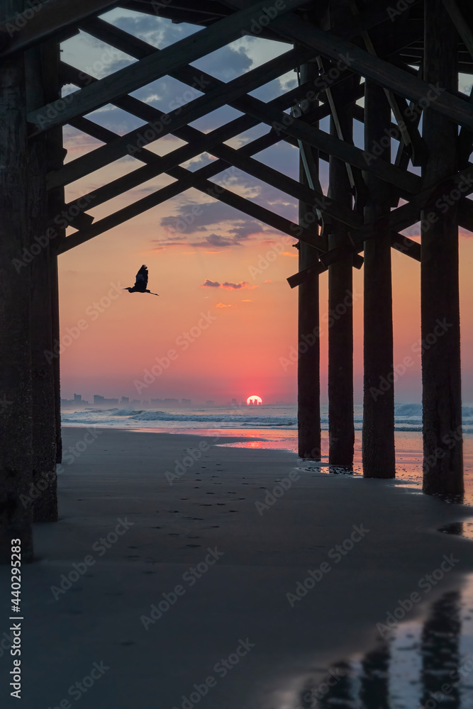 Sunrise at the beach framed by a pier with blue heron flying through