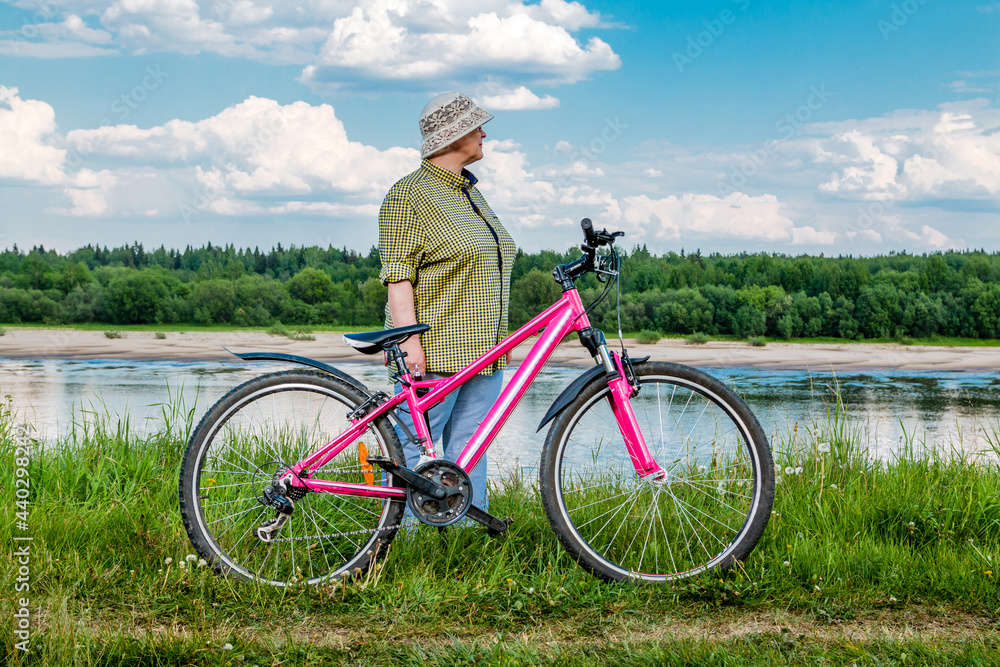 Obraz premium Elderly woman with a bicycle on the river bank. Grandma rides a bike.