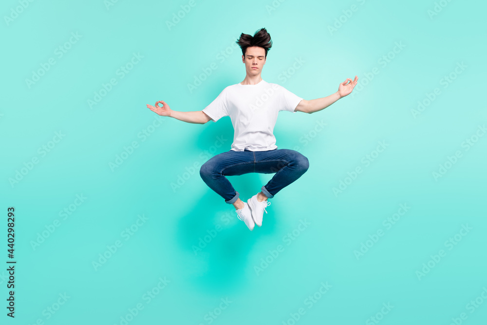 Full size photo of calm brunet hairdo teenager guy jump do yoga wear t-shirt jeans isolated on teal background
