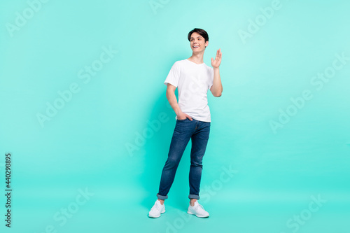 Full size photo of positive brunet hairdo teen guy wave empty space wear t-shirt jeans isolated on teal color background