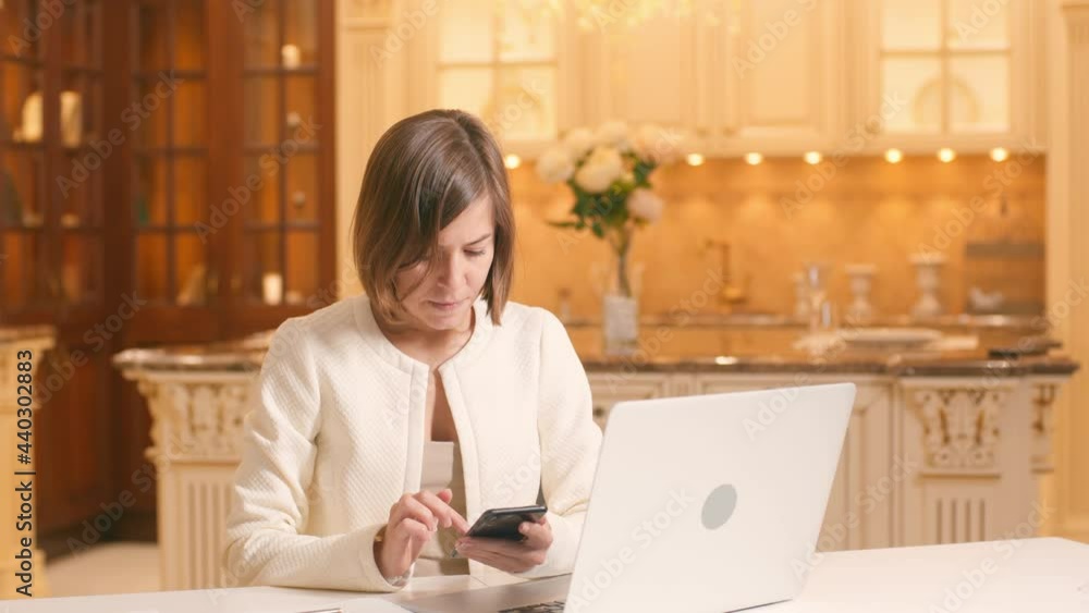 Business Woman In White Clothes Sitting At Desk In Home Office Workplace Using Smartphone Checking Information Online In Internet. Businesswoman Use Mobile Cell Phone On Domestic Kitchen Interior. Business Woman In White Clothes Sitting At Desk In Home Office Workplace Using Smartphone Checking Information Online In Internet. Businesswoman Use Mobile Cell Phone On Domestic Kitchen Interior.