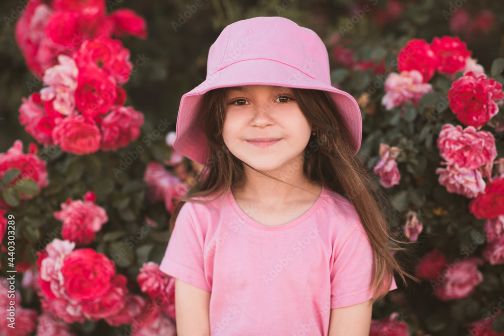 Cute child girl 5-6 year old wear hat and summer clothes posing with garden roses outdoors. Summer season. Childhood. Stylish toddler over nature background. Looking at camera. 