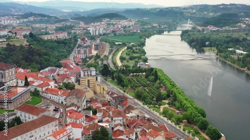 Aerial high angle pan shot over Coimbra City, green park and Mondego River in Portugal. Beautiful idyllic landscape with mountains in backdrop.