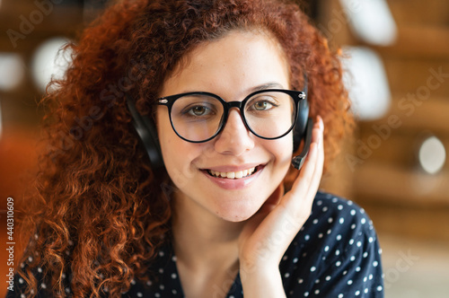 Attractive redhead curly woman wearing wireless headset and stylish eyewear looks at the camera, takes a call and smiling, close-up portrait of responsible support representative in touch, webcam view