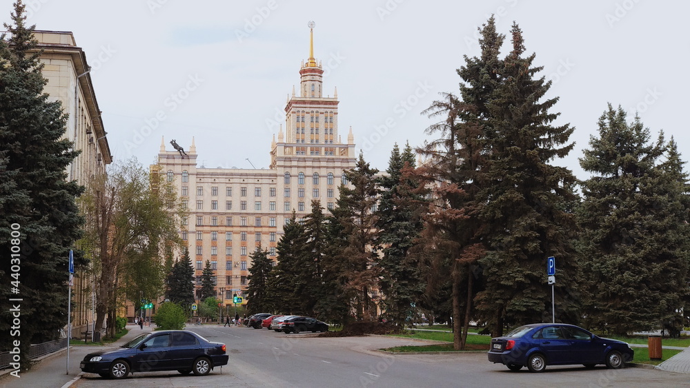 A road and autos and a large number of fir trees with a Soviet building in the background