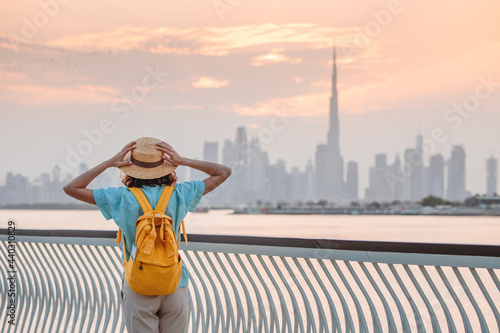 A happy traveler woman with a hat and a yellow backpack enjoys a stunning panoramic view of the Dubai Creek Canal and the famous tallest skyscraper Burj Khalifa