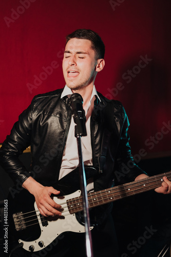 Talented handsome young guitarist man singing a song in studio recording on red background surrounded by instruments. Passion, hobby, singer, electric guitar