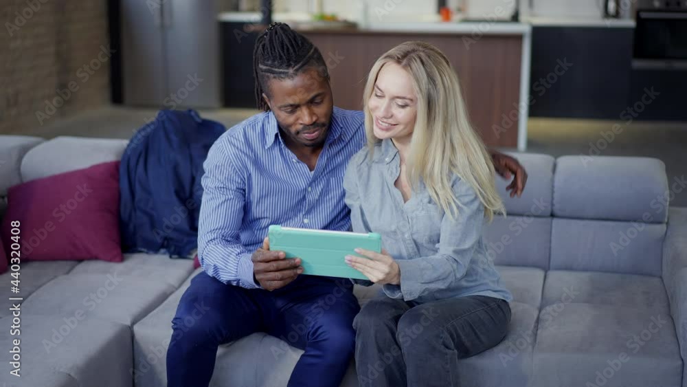 Interracial couple hugging sitting on couch surfing Internet on tablet Positive African American husband and Caucasian wife talking scrolling social media on device screen. Slow motion Interracial couple hugging sitting on couch surfing Internet on tablet Positive African American husband and Caucasian wife talking scrolling social media on device screen. Slow motion