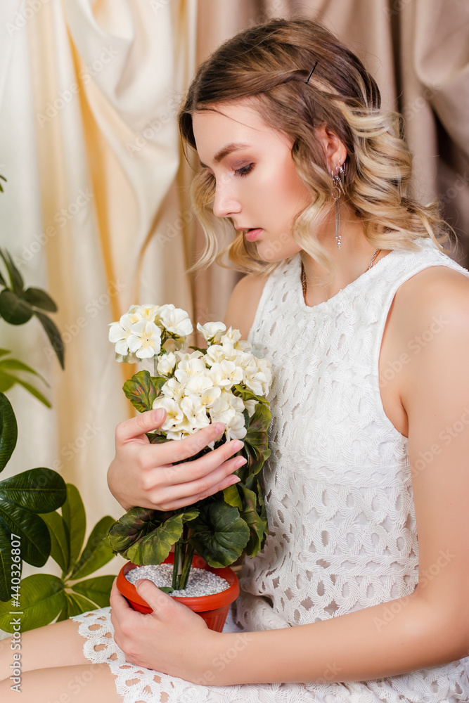 Portrait of a beautiful young woman in a white dress with a pot of artificial white flowers in her hands sitting on a chair against a background of light beige drapery.