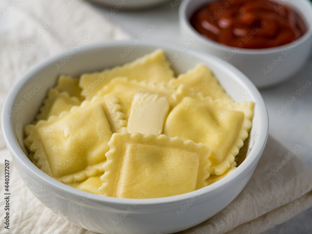 In the foreground, we see a white earthenware plate of ravioli. In the background is a deep bowl of sauce. White background. Careful viewing. High angle view. There are no people in the photo. In the foreground, we see a white earthenware plate of ravioli. In the background is a deep bowl of sauce. White background. Careful viewing. High angle view. There are no people in the photo.