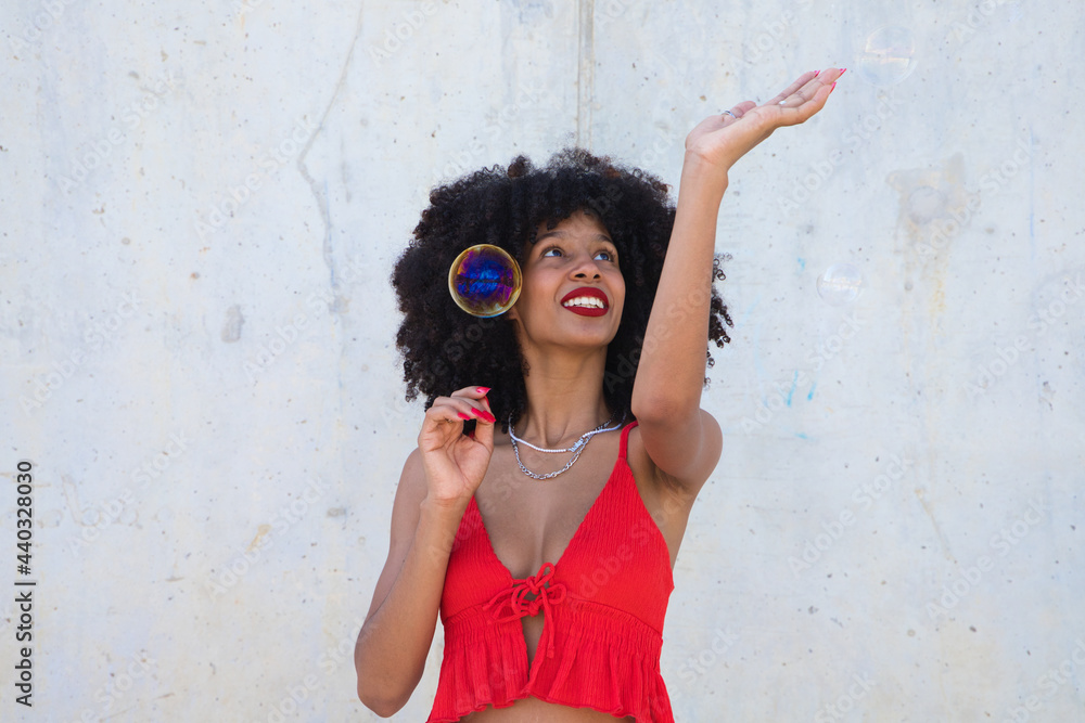 Obraz premium Beautiful afro american woman playing with soap bubbles. The woman is blowing the bubbles and hides among the soap bubbles. Happiness concept