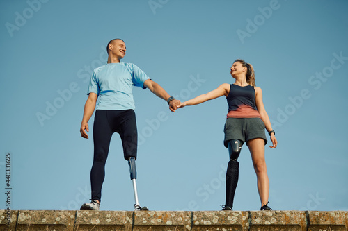 Happy handicapped couple with artificial legs holding hands and looking at each other against the sky.