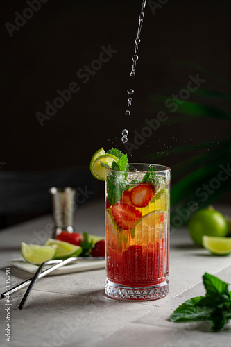 Lime, mint and strawberry refreshing cold drink - homemade lemonade or cocktail with beautiful  glass styling, droplets. Dark background with tropic plant. Recipe ingredients on the grey table.