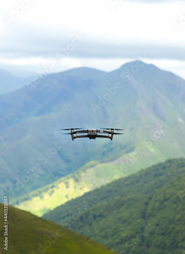 Drone flies away over a mountainous landscape. The drone can be seen from behind flying away. In the background you can see a mountain and below it a forest and a green meadow.