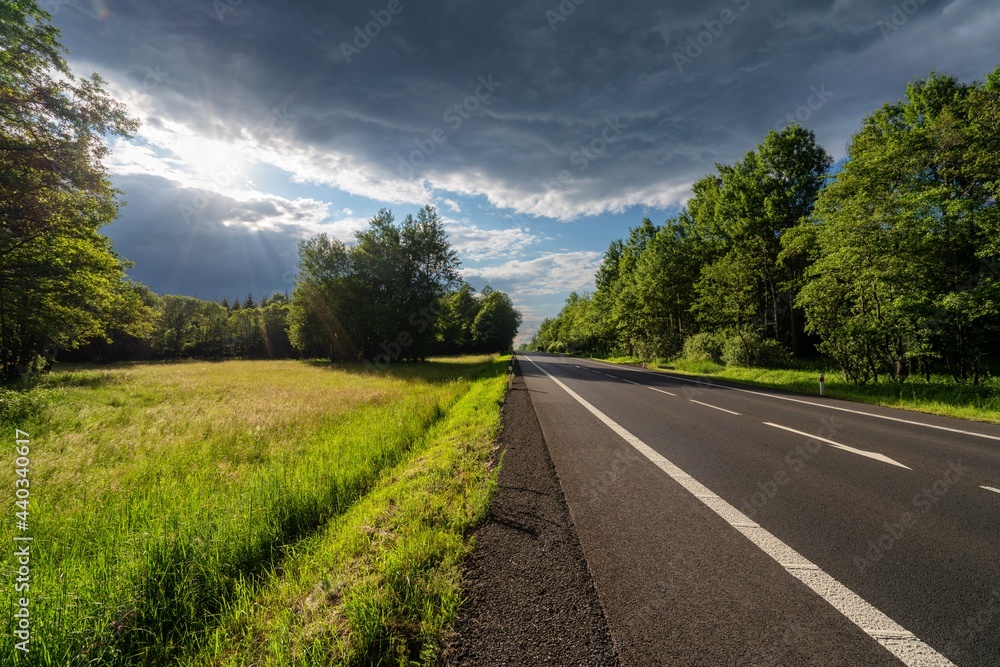 Empty asphalt road between the forest and the clearing at sunset with dark cloud