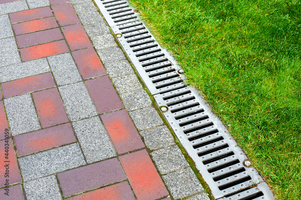 iron grating line of the drainage system of the pedestrian sidewalk made of stone tiles landscaping of the park with engineering systems on side of the path with the green lawn of the park, nobody.
