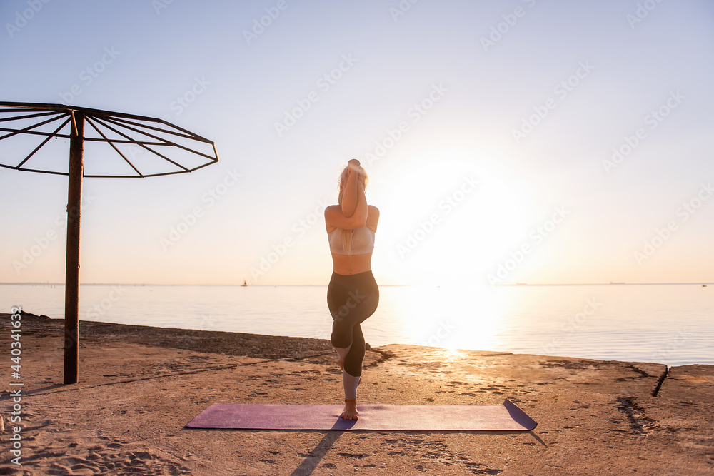 Young blonde woman in sportswear doing yoga asanas Garudasana - eagle pose  on the seashore at sunrise on a purple yoga mat. Sports activities,  meditation by the ocean on yellow sand Stock