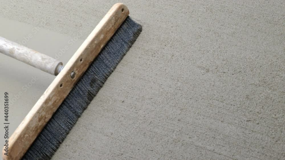 Newly poured cement concrete sidewalk is finished with a slightly rough walking surface, by the use of a wide push broom that is pulled lightly over it. Closeup clip from overhead perspective.