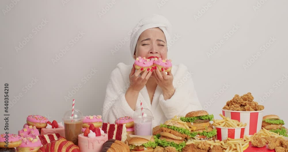 Upset young depressed Asian woman eats tasty donuts consumes much calories fast food wears white soft bathrobe and towel isolated over white background wnats to cry. No diet day. Sugar addiction