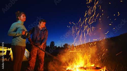 Camping couple at night. Man and woman stand by the bright bonfire with tent and car on the background