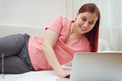 Smiling woman with red hair in pajamas working remotely on laptop while lying in bed