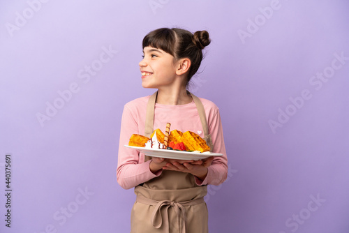 Little caucasian girl holding waffles isolated on purple background looking side