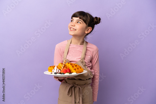 Little caucasian girl holding waffles isolated on purple background looking to the side and smiling