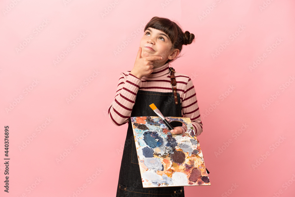 Little artist girl holding a palette isolated on pink background having doubts Little artist girl holding a palette isolated on pink background having doubts