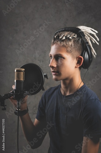 Vertical shot stilysh guy with dreadlocks is recording a song in the studio. A young attractive singer in black studio headphones stands in front of a microphone and sings. Generation Z young talent photo