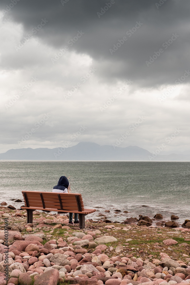 Teenager girl sitting on a bench and enjoys view on Atlantic ocean, low cloudy sky. Croagh Patrick in the background. Travel and tourism concept. Vertical image