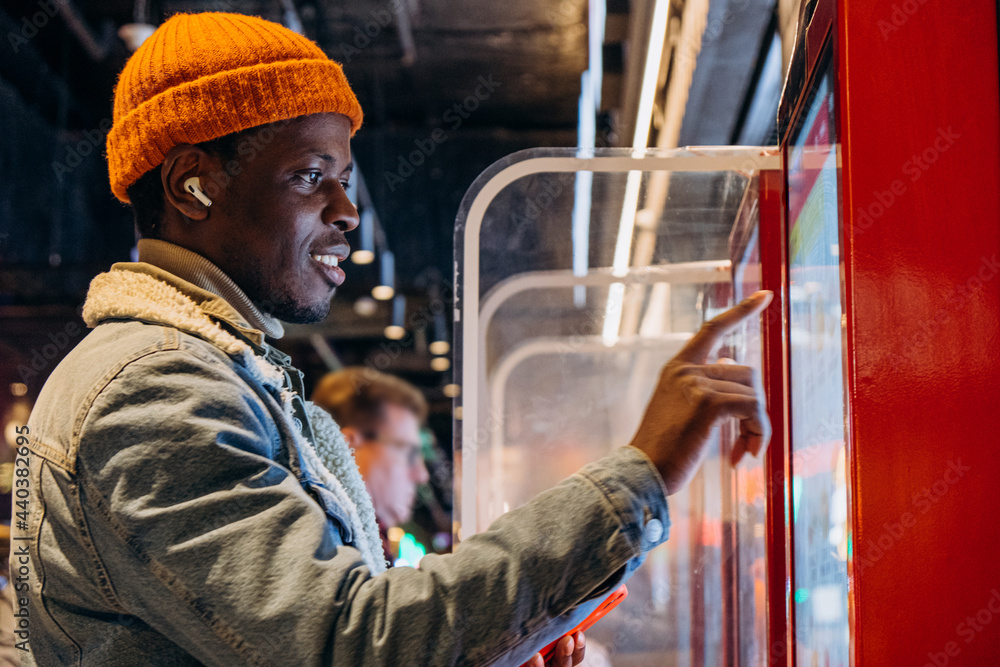 Positive African-American guy in orange knitted hat with wireless earphones orders food through self-service kiosk in cafe Positive African-American guy in orange knitted hat with wireless earphones orders food through self-service kiosk in cafe