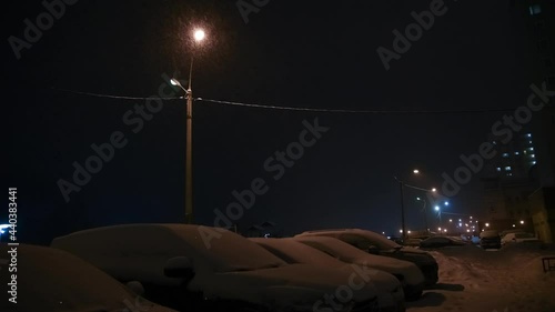 Selective focus. Heavy snow during blizzard with street lamp light at winter. Parked cars. Blurred apartment building in the background. 4K resolution video. Winter weather forecast theme.