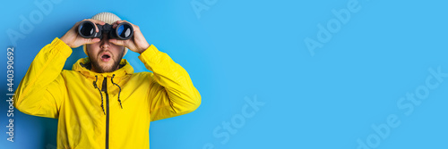 Fotografia surprised young man in a yellow jacket looks through binoculars to the side on a blue background