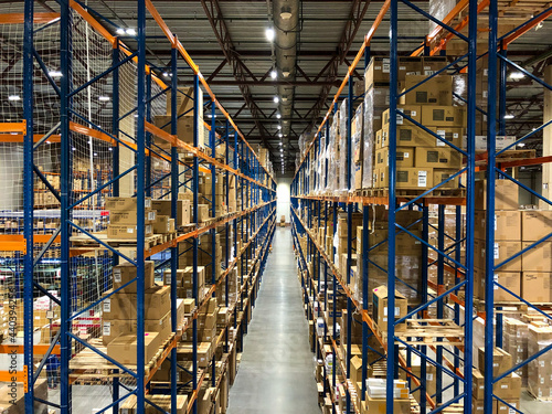 Perspective view to the warehouse, blue metal racks, shelves full of goods packed in carton boxes or pallets. Clean and tydy distribution center. 