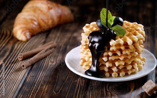 Pouring melted chocolate on a stack of freshly baked belgian waffles decorated with blueberry and mint on white plate. Delicious homemade pastry for breakfast on wooden table or rustic background