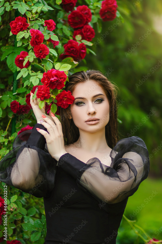 Beautiful girl in the garden with roses. Portrait of a woman with big lips in black clothes on a background of red flowers. Greens
