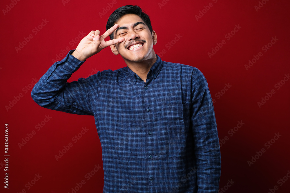 Young handsome asian man wearing casual shirt doing peace symbol with fingers over face, smiling cheerful showing victory